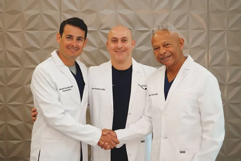 Three male dentists in white lab coats standing side-by-side smiling, with two shaking hands in front of a textured wall background