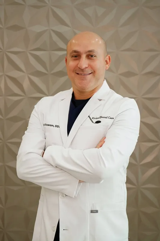 Professional headshot of a bald male dentist with arms crossed, smiling in a white lab coat over a dark shirt