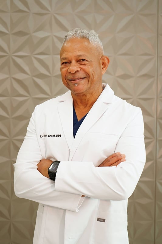 Professional headshot of an older male dentist with short grey hair smiling with arms crossed in a white lab coat over dark scrubs