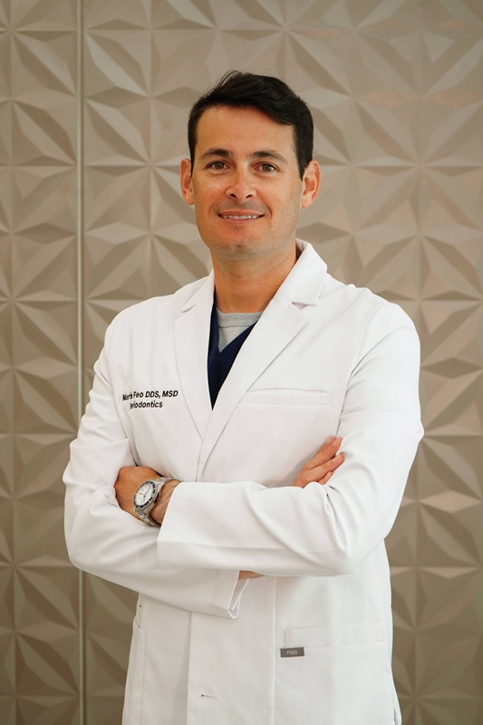 Professional headshot of a male dentist with dark hair, smiling with arms crossed in a white lab coat over dark scrubs
