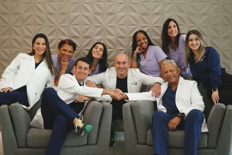 Group photo of a dental team of nine healthcare professionals smiling together in a modern clinic with grey patterned wall decor