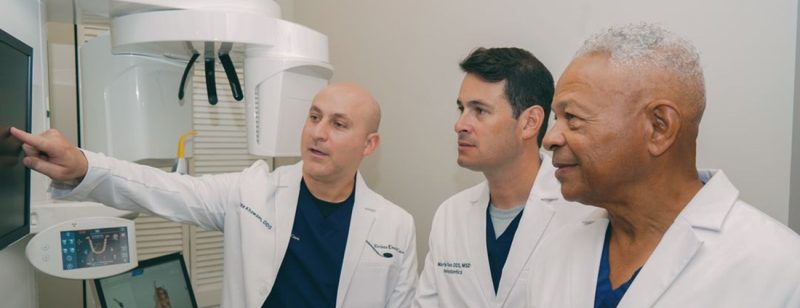 Three male dentists reviewing digital X-ray scans together on a monitor screen inside a modern dental treatment room