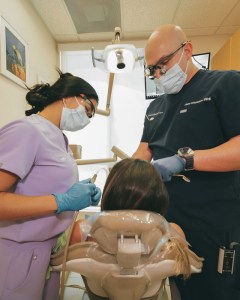A dentist wearing a face mask and protective glasses performing a procedure on a patient alongside an assistant