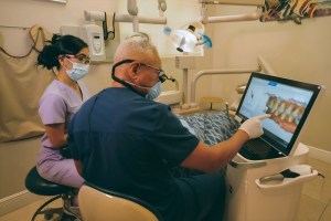 A dentist pointing at a 3D digital dental scan on a computer monitor while discussing treatment with an assistant in an operatory room